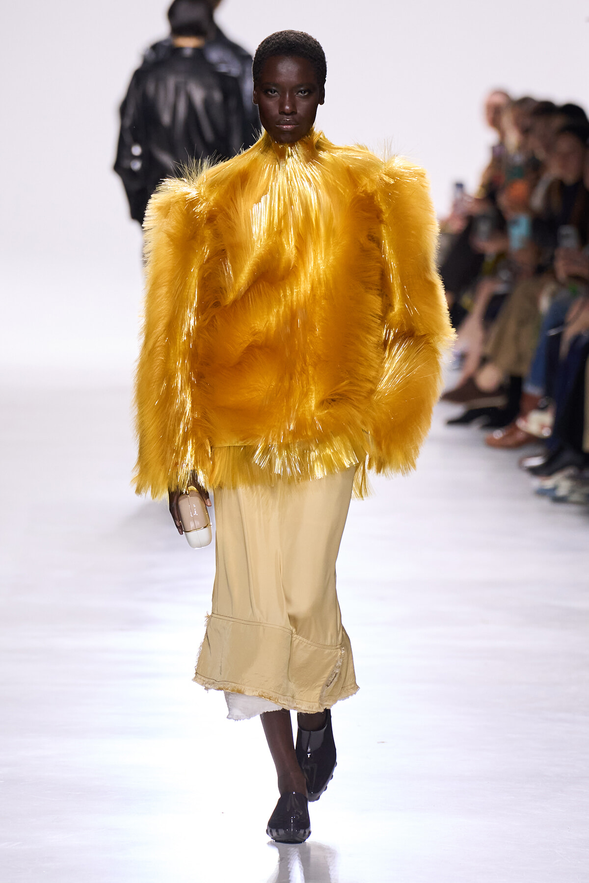 Model on a runway wearing a bright yellow shaggy fur coat over a beige midi skirt, carrying a small clutch.