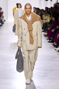 Male model on a runway wearing a beige double-breasted suit with a camel scarf draped over the shoulders and a gray woven tote by his side