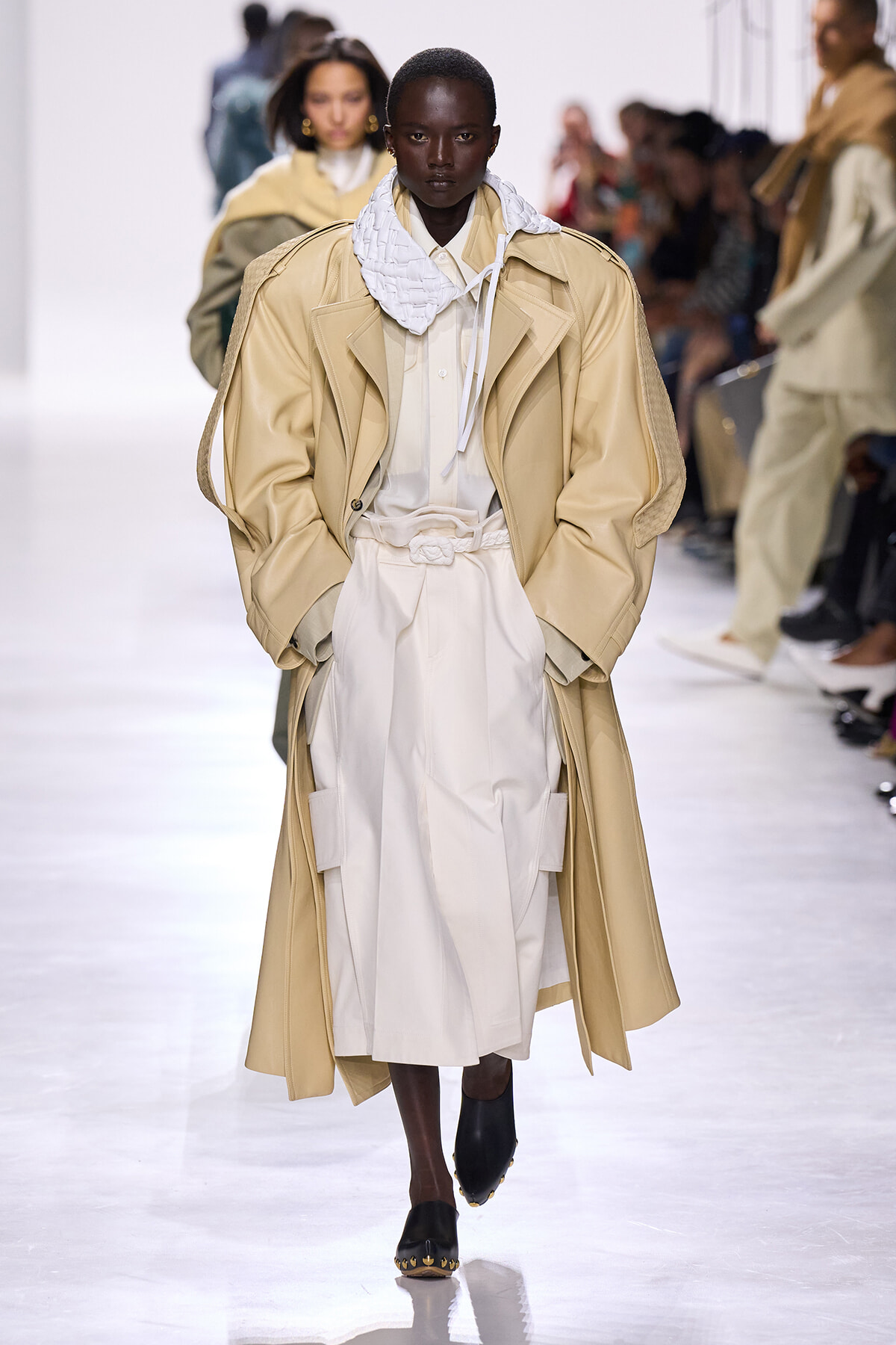 Model walking the runway in a beige oversized trench over a white outfit with a chunky white scarf, black loafers, and minimalist accessories.
