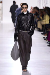 Female model on a runway wearing a black leather jacket, textured dark pants, large gray woven bag, and sunglasses.