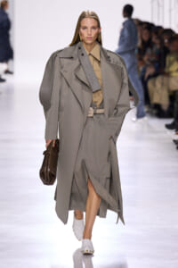 Model walks a runway in an oversized gray trench with braided shoulder detail, tan shirt, and beige belt, carrying a brown textured clutch.