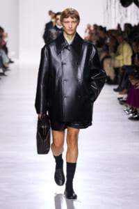 Model on a fashion runway wearing an oversized black glossy leather coat, black shorts, and matching bag, stepping forward confidently.