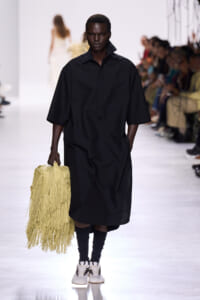 Model walking a fashion runway in a loose black dress, carrying a yellow fringe handbag with a crowd in the background