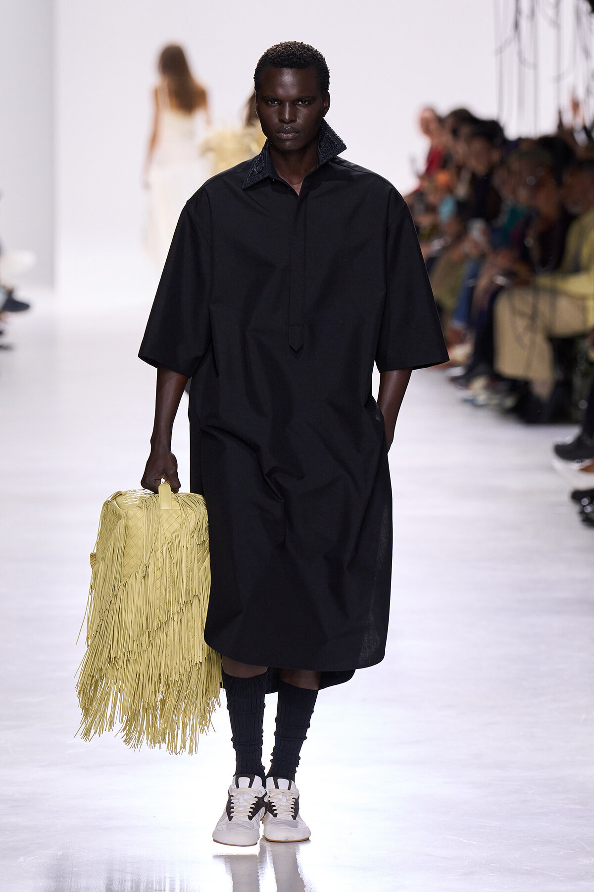 Model walking a fashion runway in a loose black dress, carrying a yellow fringe handbag with a crowd in the background