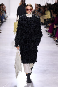 Model walks the runway in a black shaggy coat over a white turtleneck, wearing red sunglasses and gold hoops while carrying a white fringed bag.