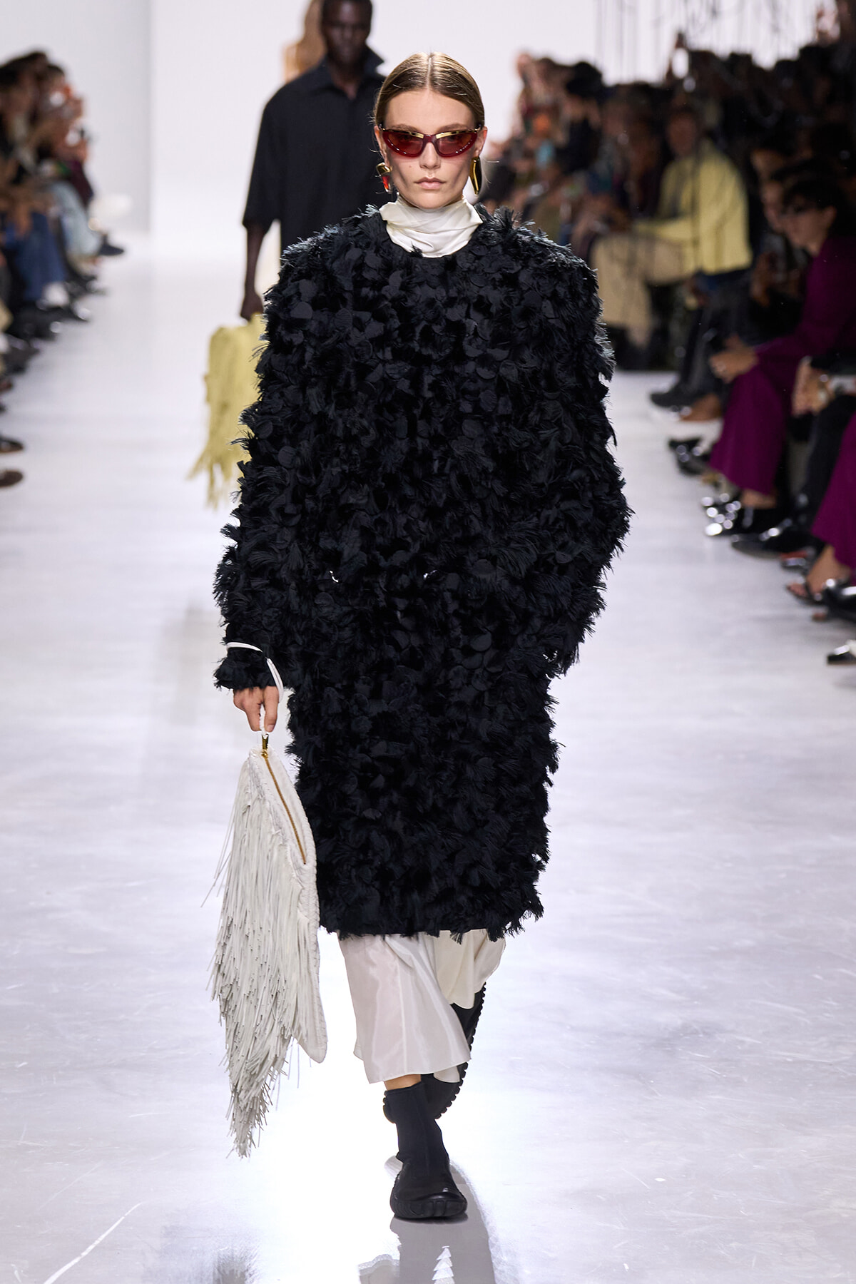 Model walks the runway in a black shaggy coat over a white turtleneck, wearing red sunglasses and gold hoops while carrying a white fringed bag.