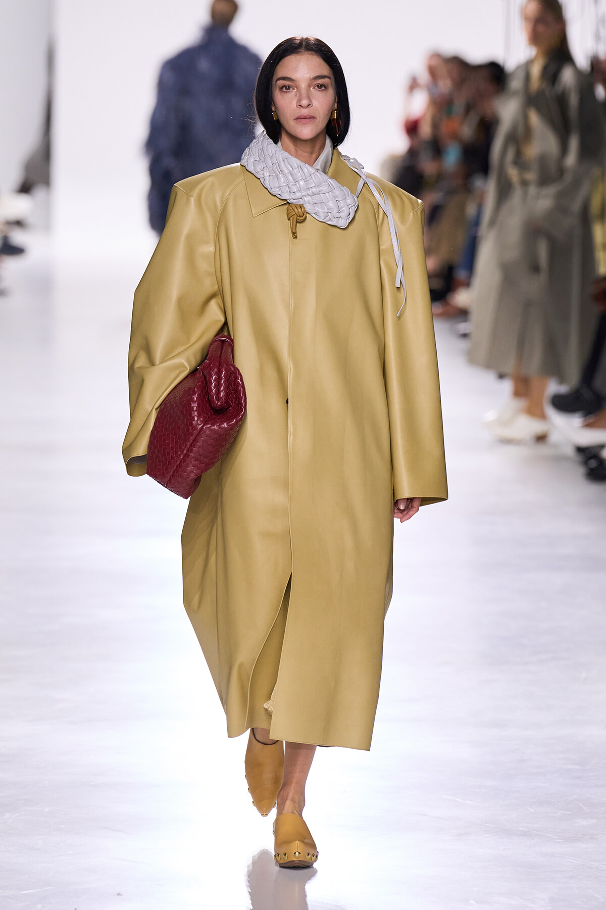 Female model on a runway wearing a long tan coat, a chunky gray scarf, mustard shoes, and carrying a red quilted clutchbag.