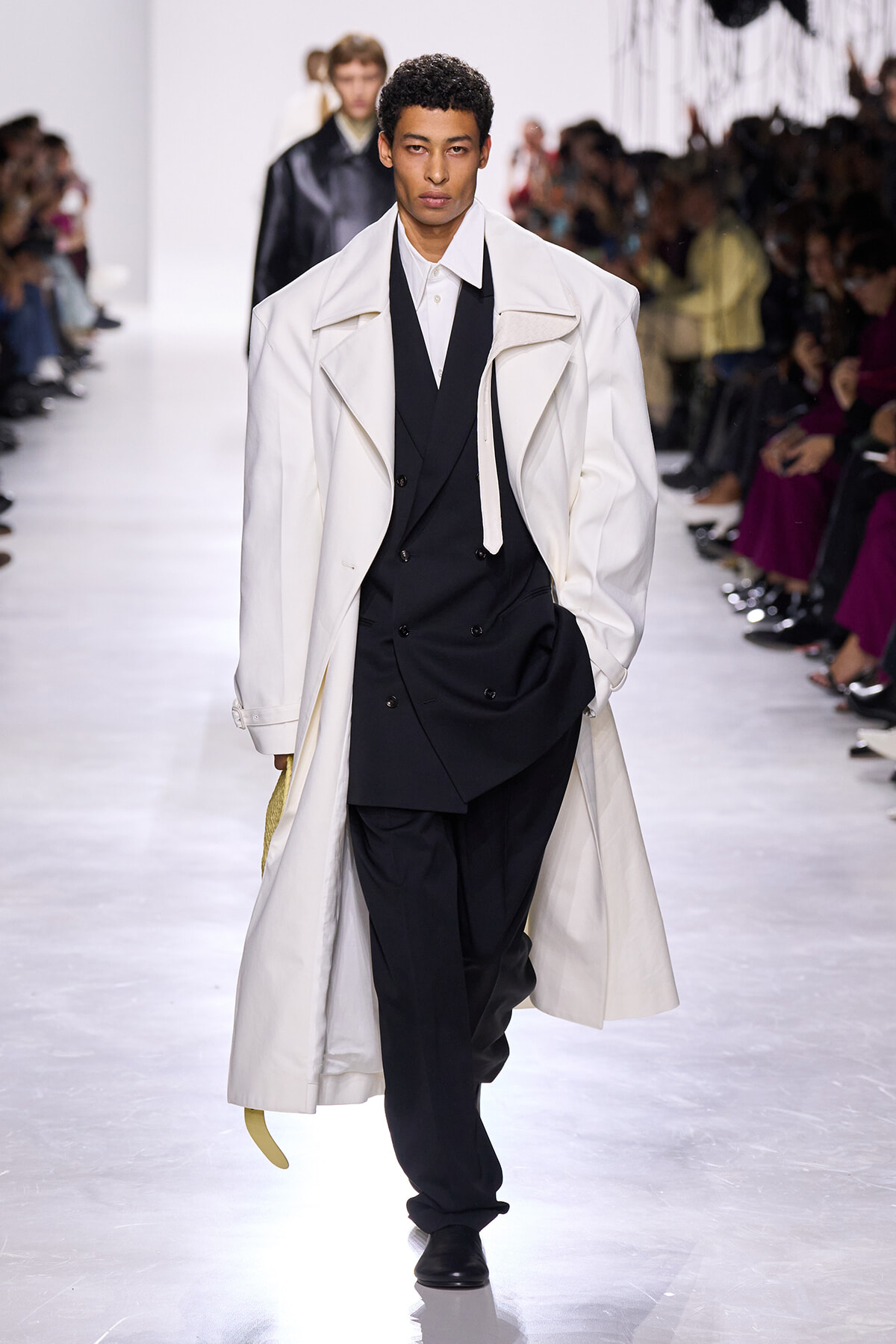 Male model walks a fashion runway wearing an oversized white trench over a black tailored suit and white shirt, with hands in pockets and audience in background.