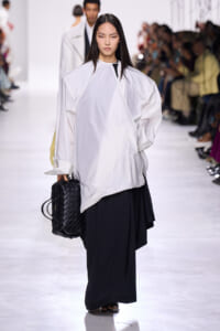 Model walking the runway in an oversized white blouse and wide-leg black pants, carrying a black quilted handbag; audience blurred in the background.