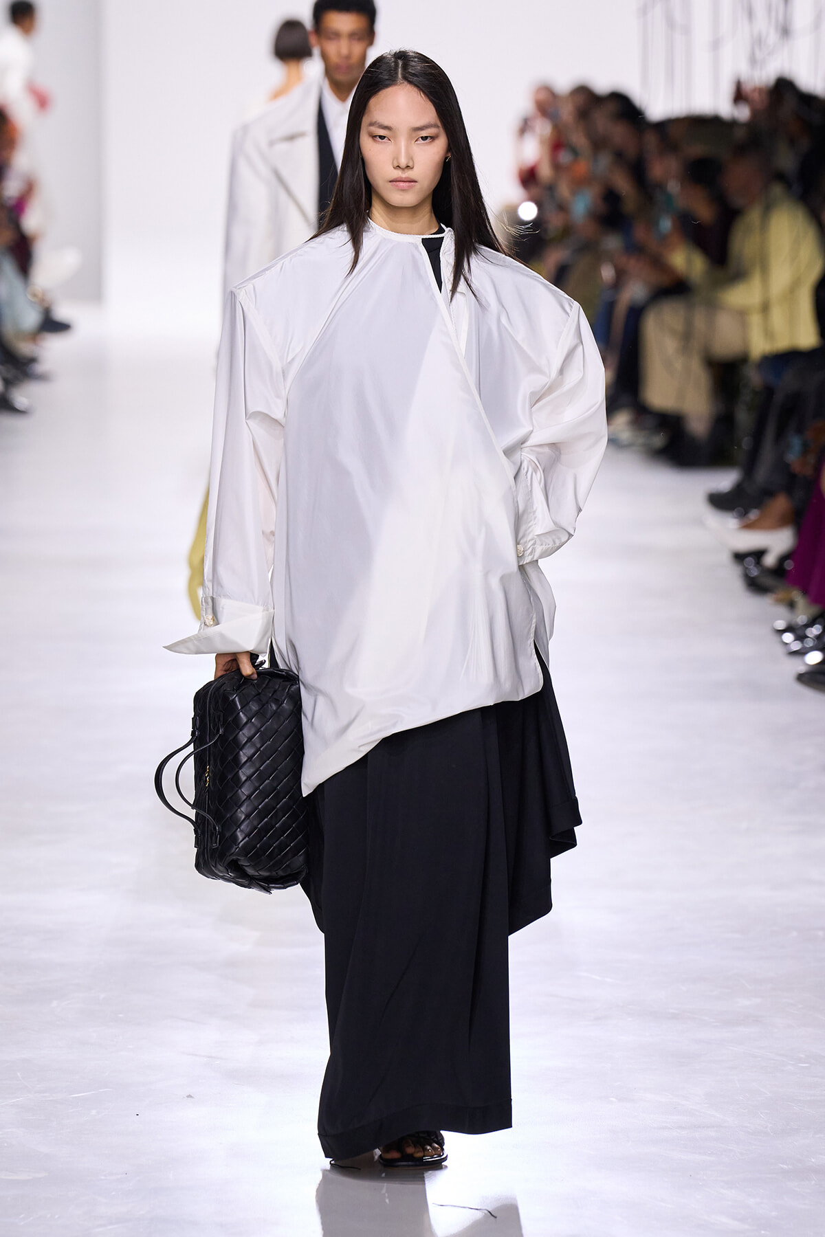 Model walking the runway in an oversized white blouse and wide-leg black pants, carrying a black quilted handbag; audience blurred in the background.