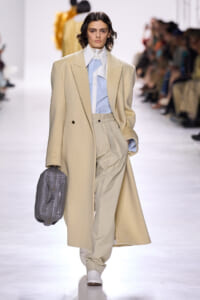 Model on a runway wearing a long beige coat over a pale blue shirt and beige pleated trousers, carrying a gray quilted backpack