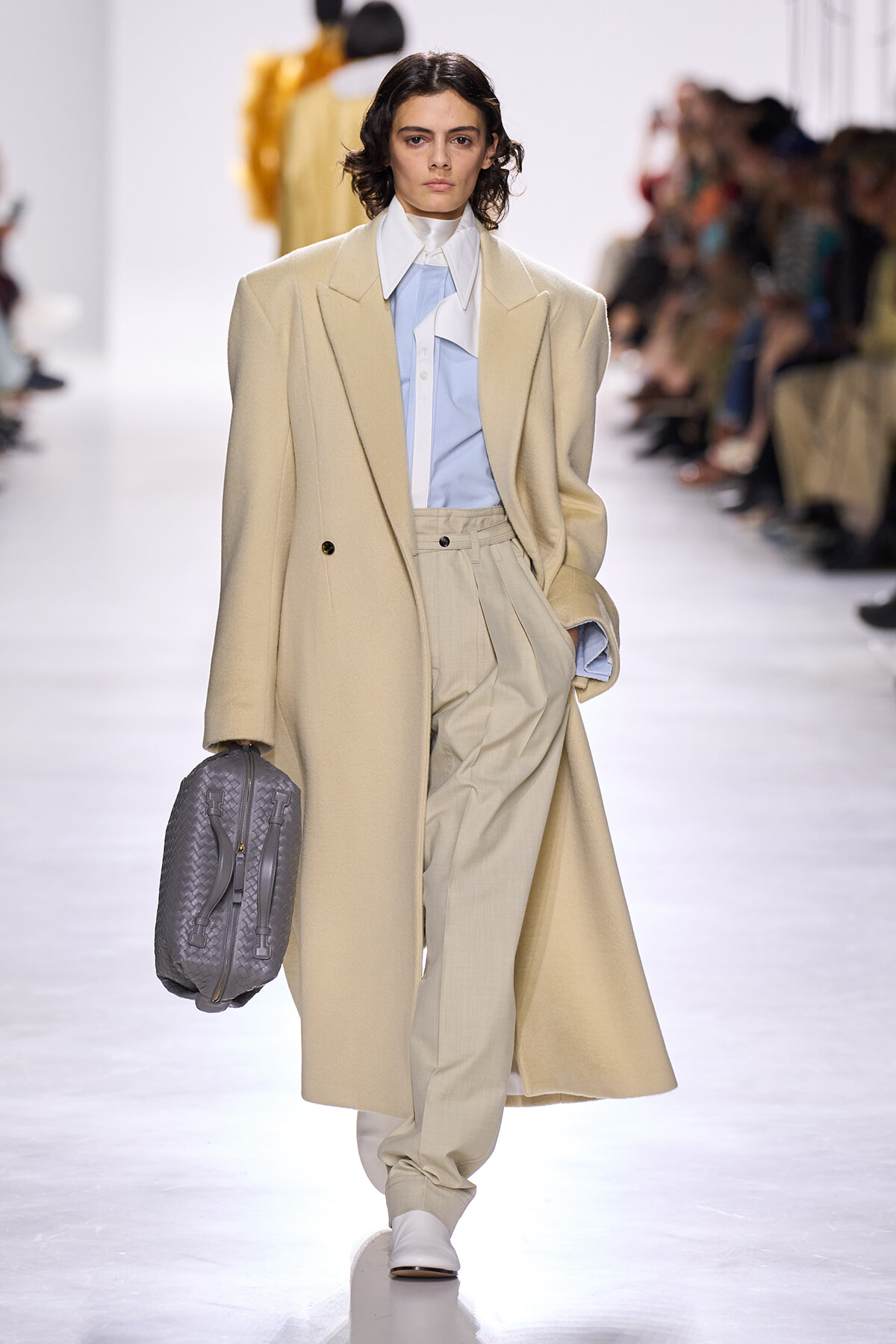 Model on a runway wearing a long beige coat over a pale blue shirt and beige pleated trousers, carrying a gray quilted backpack