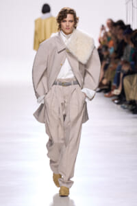 Runway model in an oversized beige suit with a white shirt and a cream fur scarf draped over one shoulder.