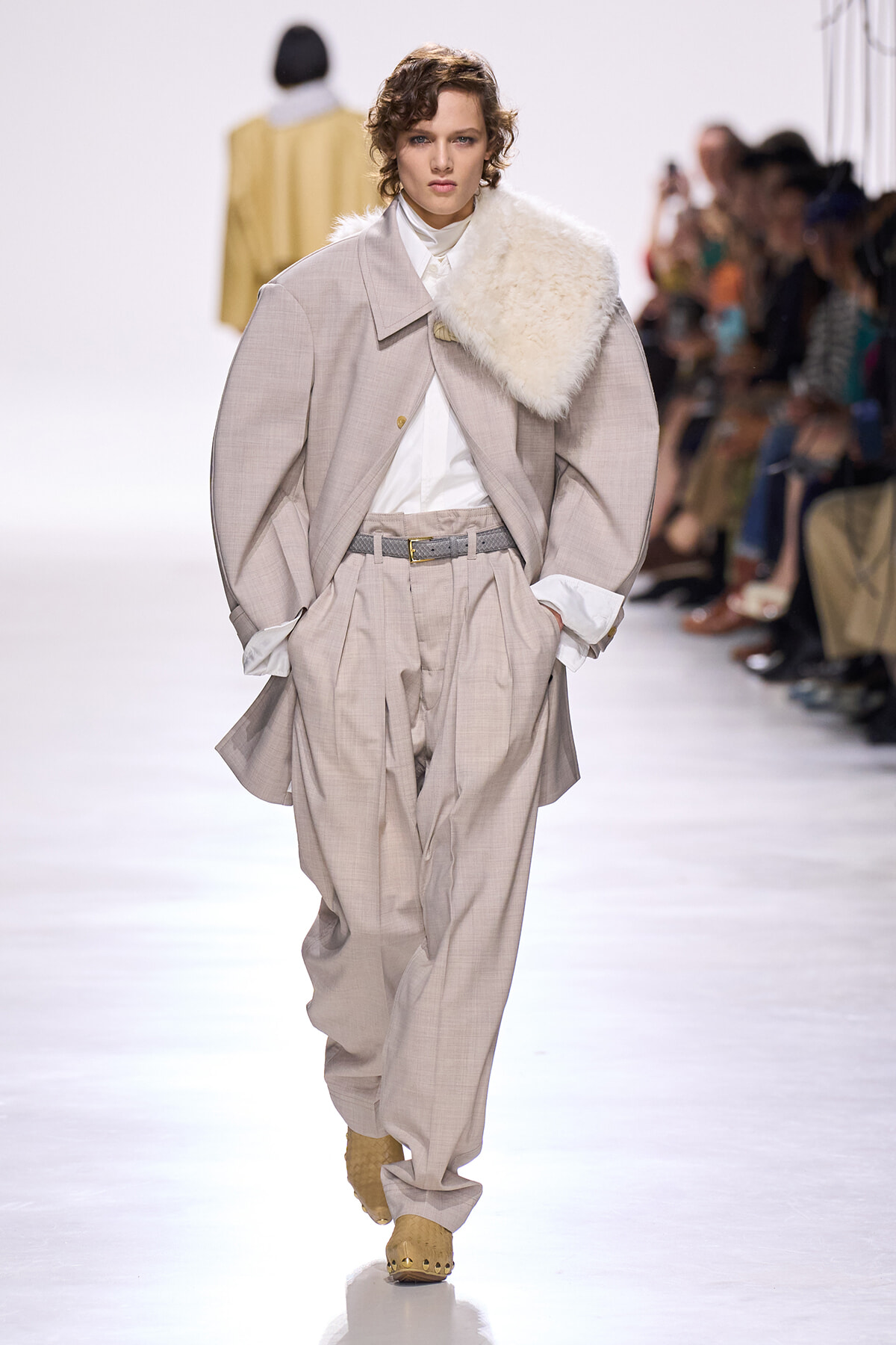 Runway model in an oversized beige suit with a white shirt and a cream fur scarf draped over one shoulder.