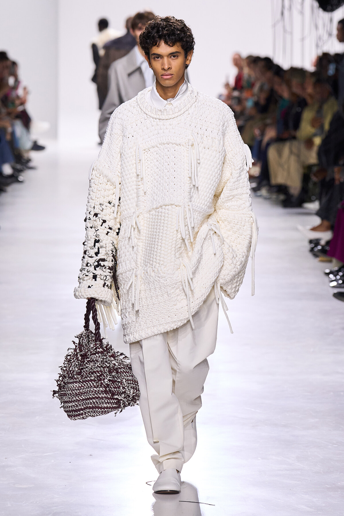 Model walks a runway in an oversized white knit sweater with fringe and tassels, white pants, and a large woven handbag.