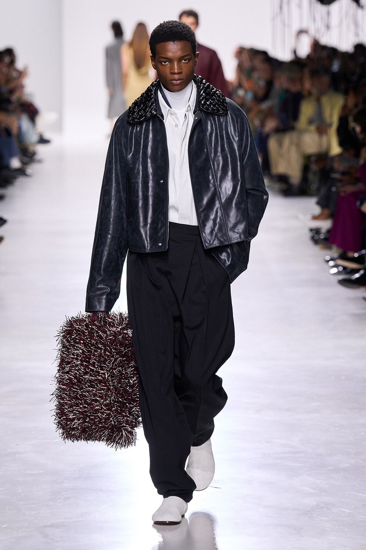Male model walking a fashion runway in a metallic navy jacket, white turtleneck, and drop-front trousers, carrying a spiky red bag.