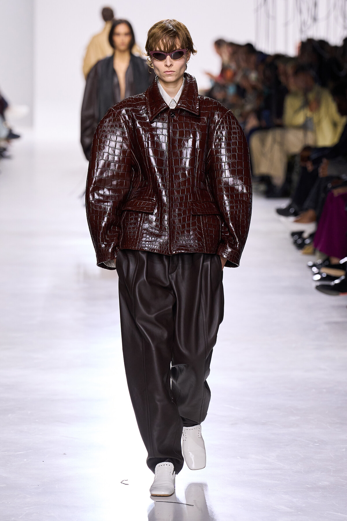 Model on a catwalk wearing an oversized burgundy crocodile-embossed jacket, black leather pants, white shoes, and purple sunglasses at a fashion show.