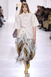 Model walks the runway in a cream oversized button-front shirt and a multi-toned feather fringe skirt.