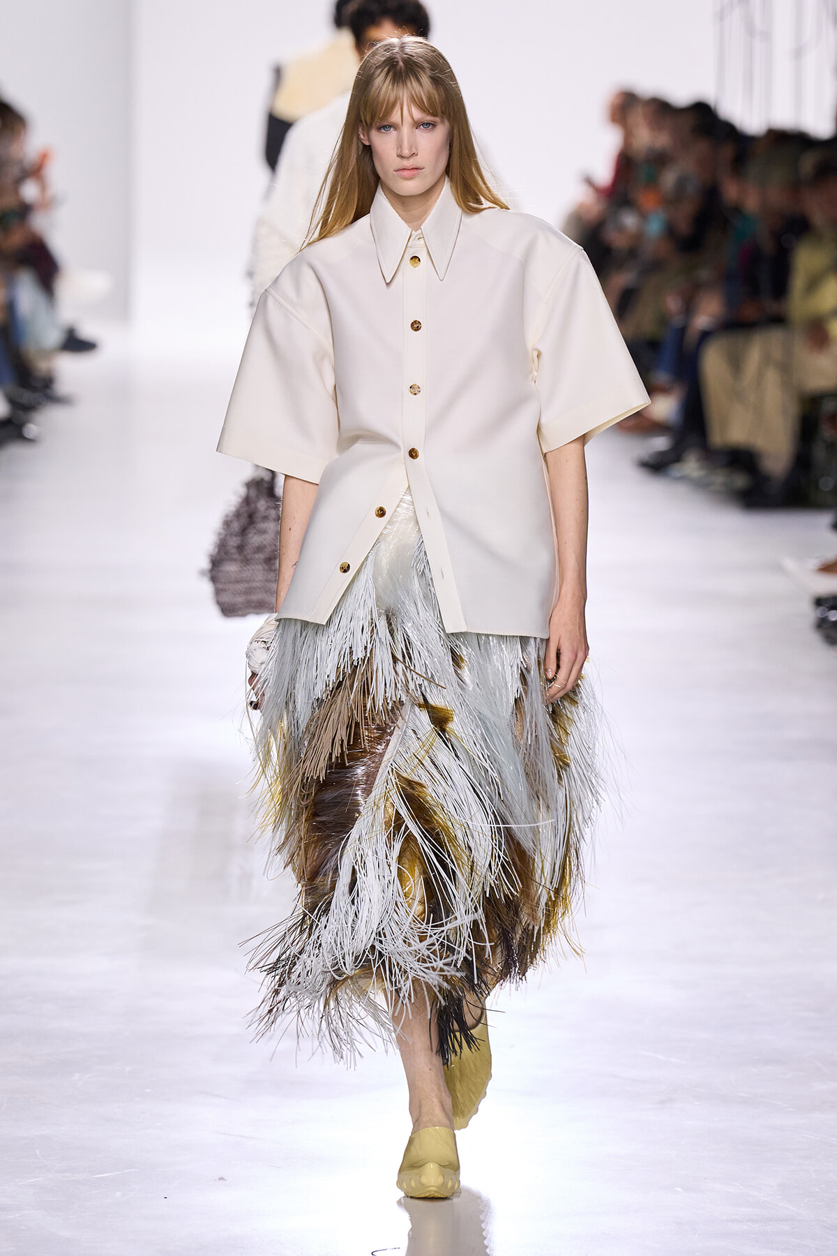 Model walks the runway in a cream oversized button-front shirt and a multi-toned feather fringe skirt.