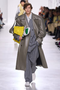 Male model walks a runway in a long gray trench over a pinstripe suit, clutching a bright yellow woven bag and papers.