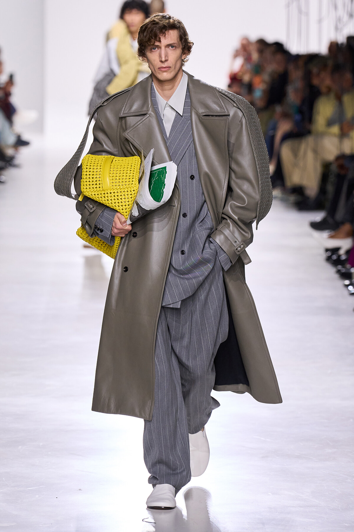 Male model walks a runway in a long gray trench over a pinstripe suit, clutching a bright yellow woven bag and papers.