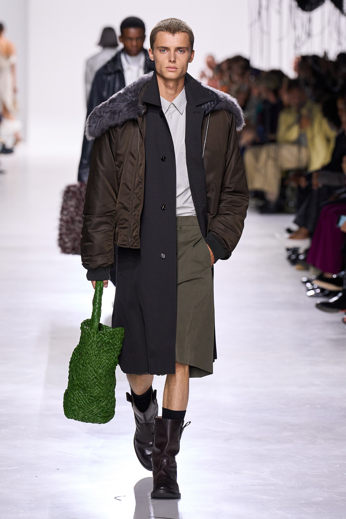 Male model walking a fashion show in a long dark coat with a fur-trim collar, olive shorts, and black boots, carrying a bright green woven bag.