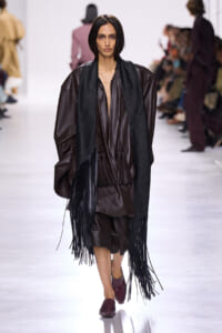 Runway model in a loose brown leather outfit with a long black fringe scarf and burgundy shoes on a white catwalk.