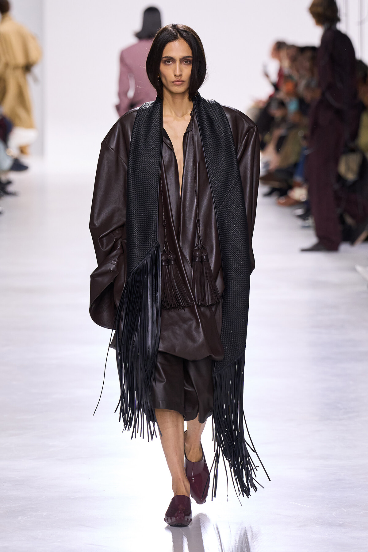 Runway model in a loose brown leather outfit with a long black fringe scarf and burgundy shoes on a white catwalk.