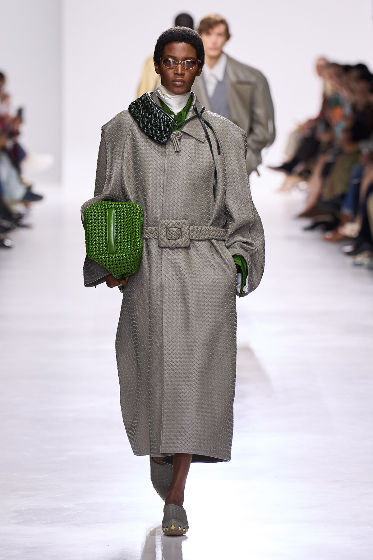 Model walks the runway in a oversized, quilted gray trench with a belt, carrying a bright green clutch and green accents.