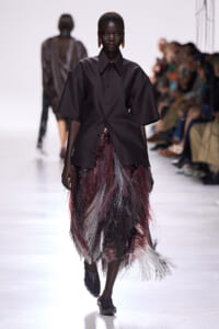 Black model walks the runway in an oversized dark shirt paired with a multi‑colored fringe skirt in burgundy, grey, and black; blurred audience visible to the sides.