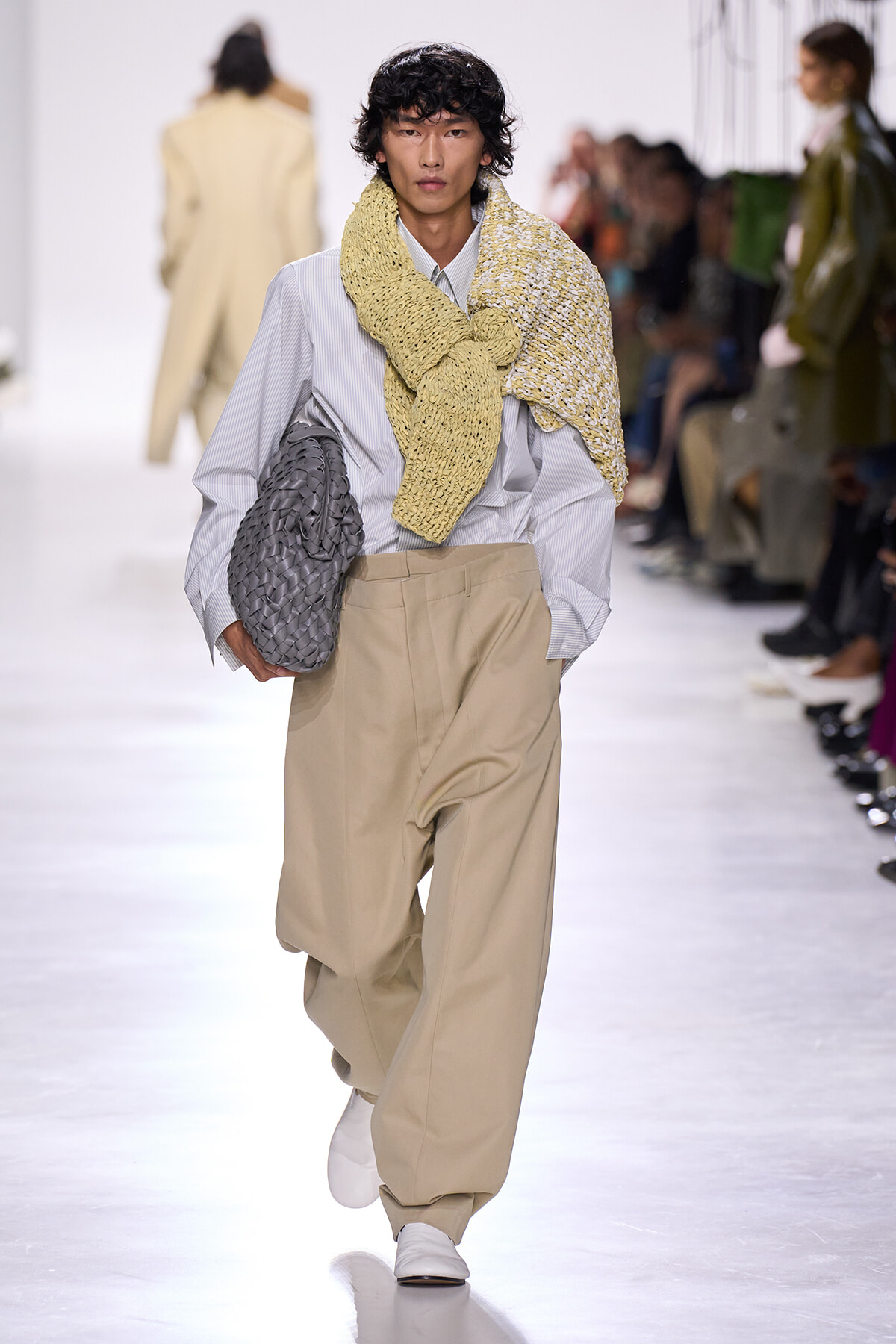 Male model walks a fashion show runway in oversized beige trousers, a pale striped shirt, a chunky yellow scarf draped over shoulders, and a gray woven bag tucked under his arm.