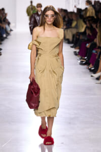 Fashion model walking a runway in a beige off-shoulder dress, carrying a red bag and wearing red fuzzy slides and white sunglasses.