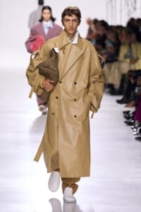 Male model walking a fashion runway in a tan long trench coat, holding a woven brown bag under his arm.