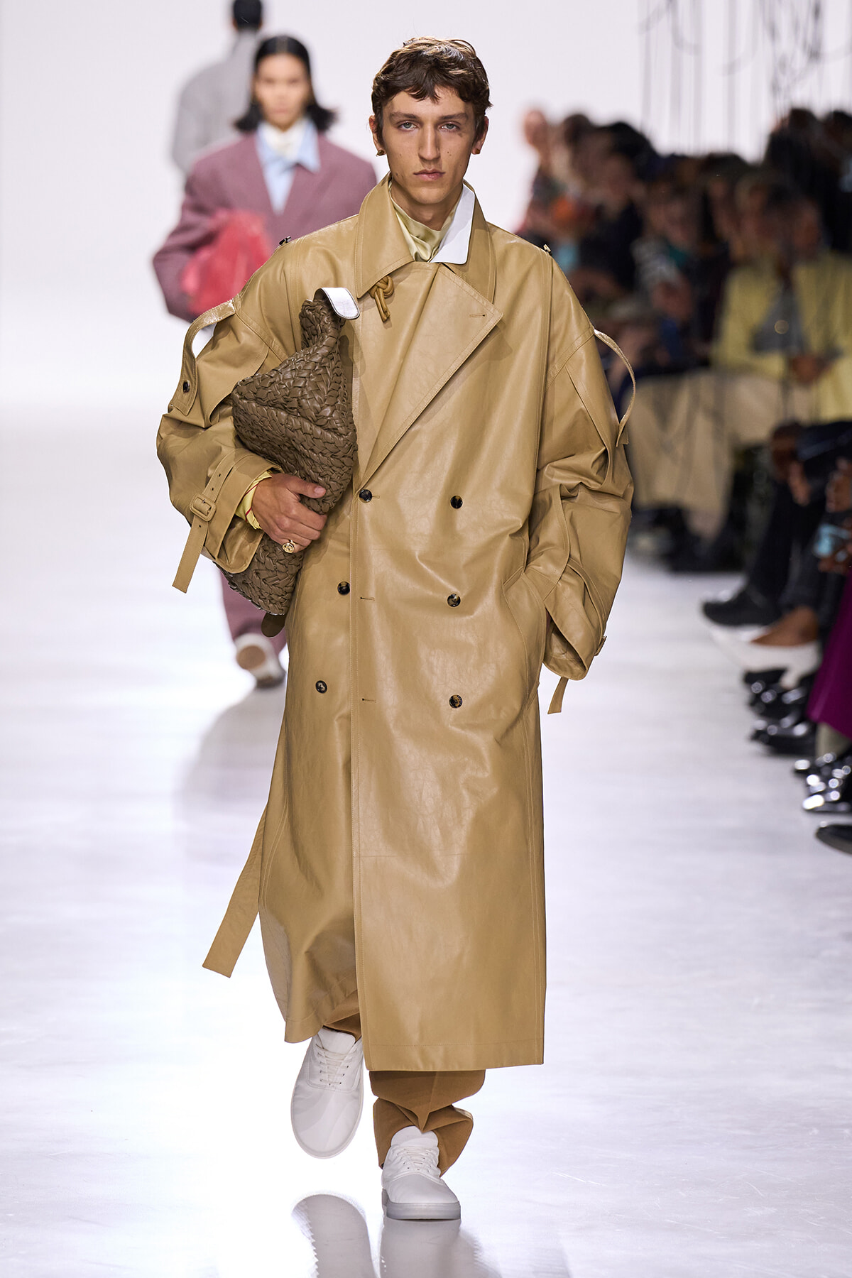 Male model walking a fashion runway in a tan long trench coat, holding a woven brown bag under his arm.