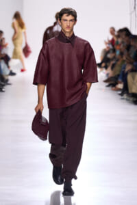 Male model in burgundy textured top and loose burgundy pants walking a runway, carrying a matching burgundy woven clutch bag.