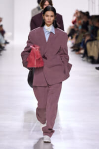 Model walks the runway in an oversized mauve suit with a light blue shirt, holding a pink fur clutch.