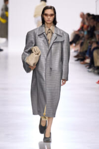 Model on a runway wearing a long gray crocodile-pattern overcoat, beige shirt, and carries a cream quilted bag; she wears tinted sunglasses.