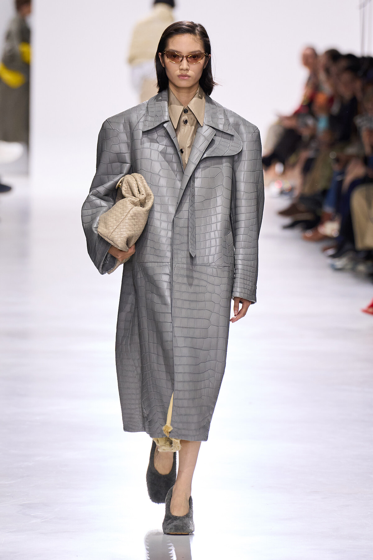 Model on a runway wearing a long gray crocodile-pattern overcoat, beige shirt, and carries a cream quilted bag; she wears tinted sunglasses.