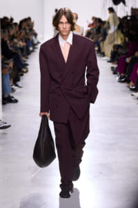 Male model on a fashion runway wearing a burgundy double-breasted suit, carrying a large black woven bag.