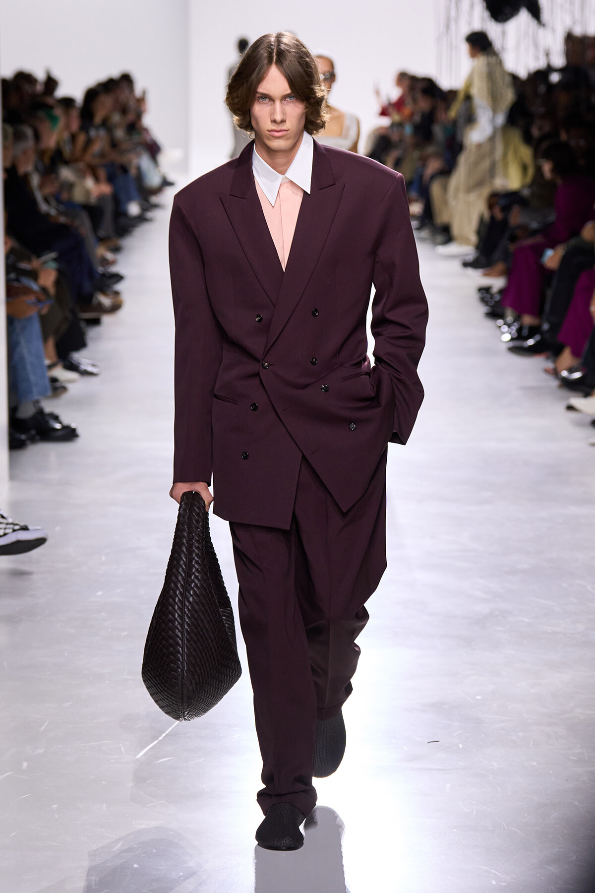 Male model on a fashion runway wearing a burgundy double-breasted suit, carrying a large black woven bag.
