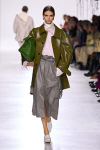 Model walking runway in olive-green leather coat over pink button blouse and gray midi skirt, holding a green textured clutch.