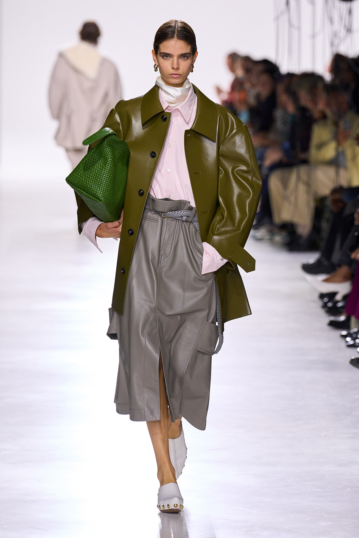 Model walking runway in olive-green leather coat over pink button blouse and gray midi skirt, holding a green textured clutch.