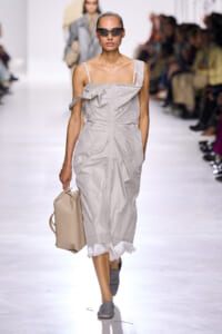 Model walks a runway in a light gray, ruched off-shoulder dress, carrying a beige tote and wearing dark sunglasses.
