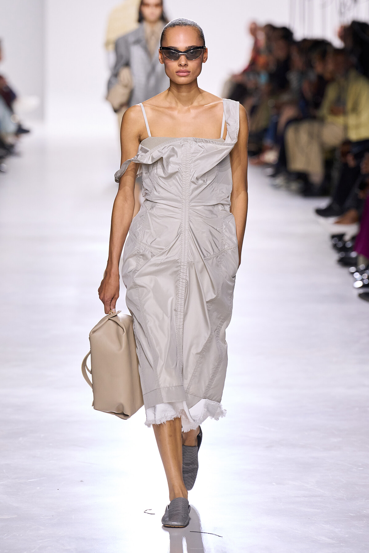 Model walks a runway in a light gray, ruched off-shoulder dress, carrying a beige tote and wearing dark sunglasses.