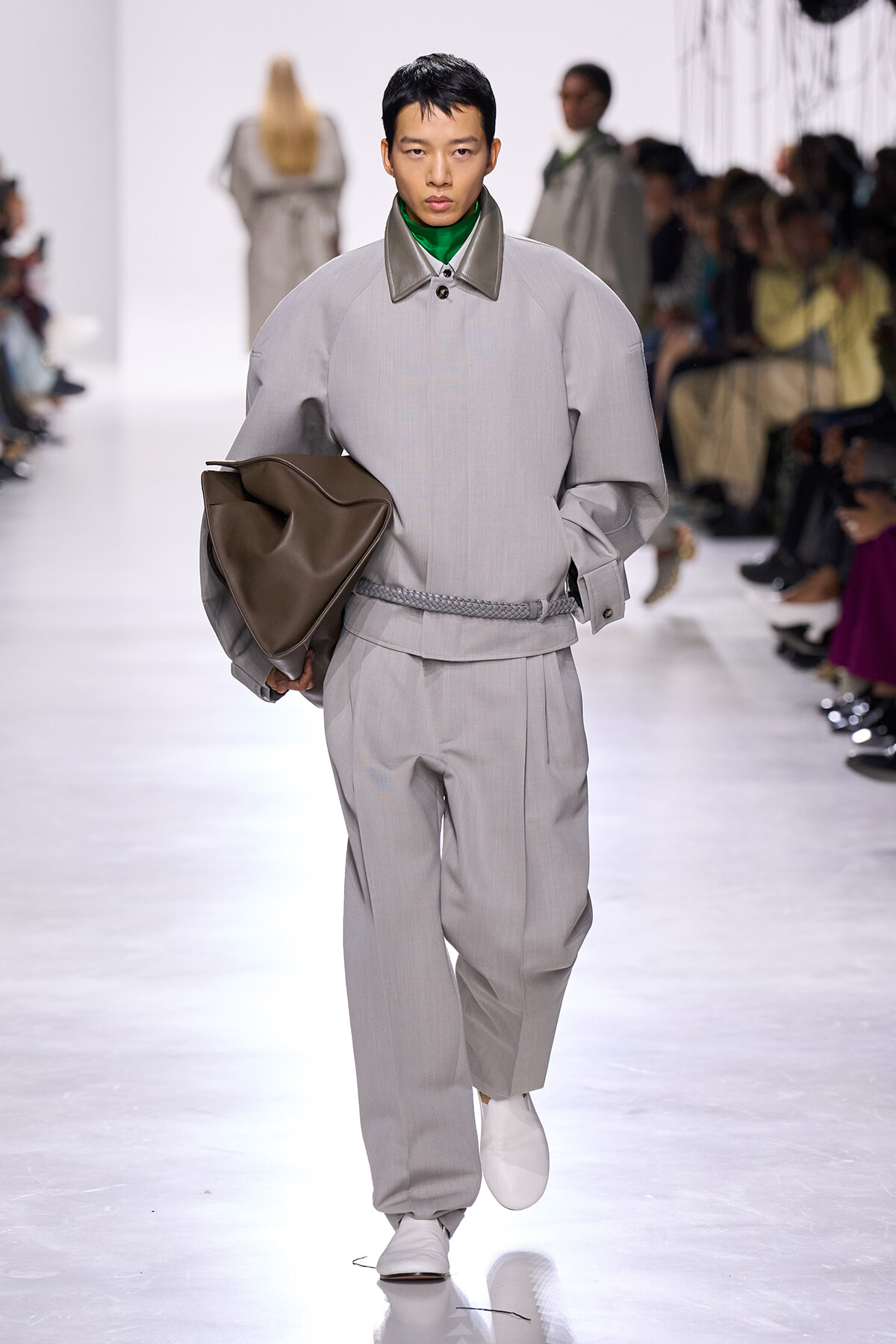 Male model walks the runway in an oversized light-gray suit with a braided belt, holding a large brown leather bag.