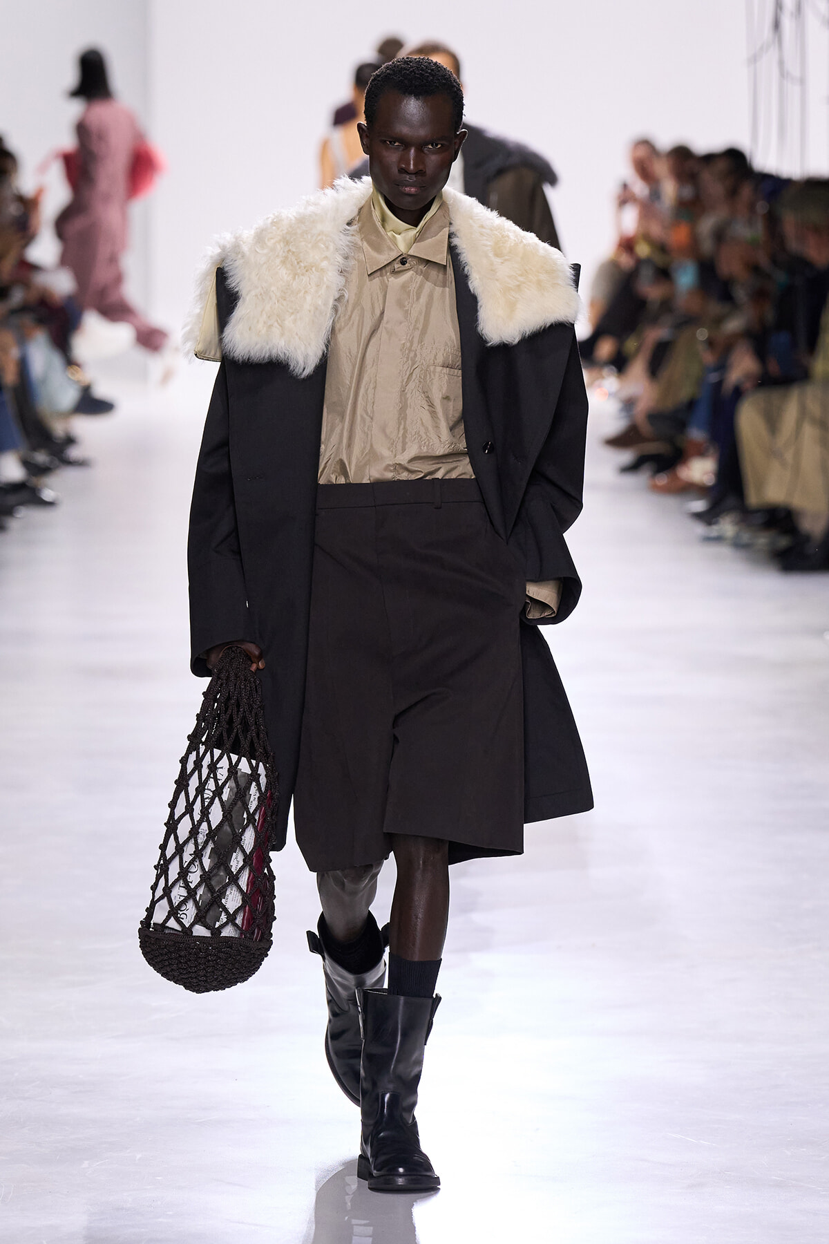Model walks a fashion runway in a black coat with a white fur collar, beige shirt, brown shorts, and black boots, carrying a black net bag.