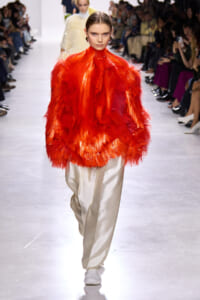Model walks the runway in a vivid red feathered oversized coat paired with cream satin pants; audience watches from both sides.