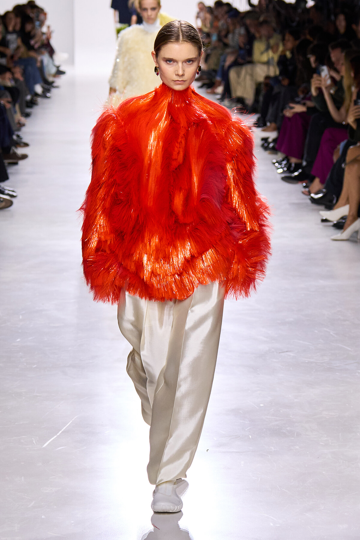 Model walks the runway in a vivid red feathered oversized coat paired with cream satin pants; audience watches from both sides.