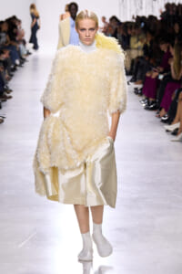 Model on a fashion runway wearing a cream shaggy sweater over a pale satin skirt with white boots.