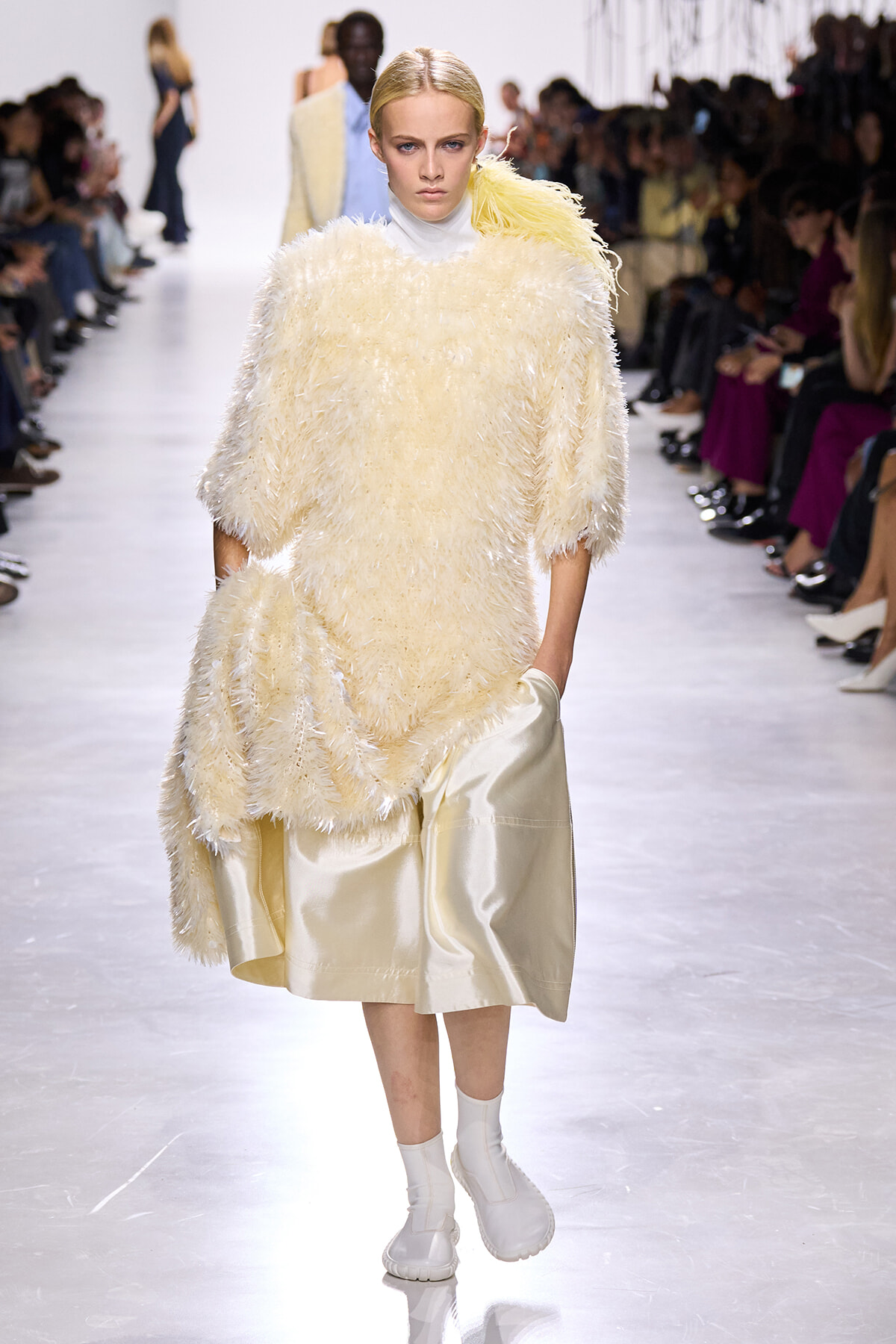 Model on a fashion runway wearing a cream shaggy sweater over a pale satin skirt with white boots.